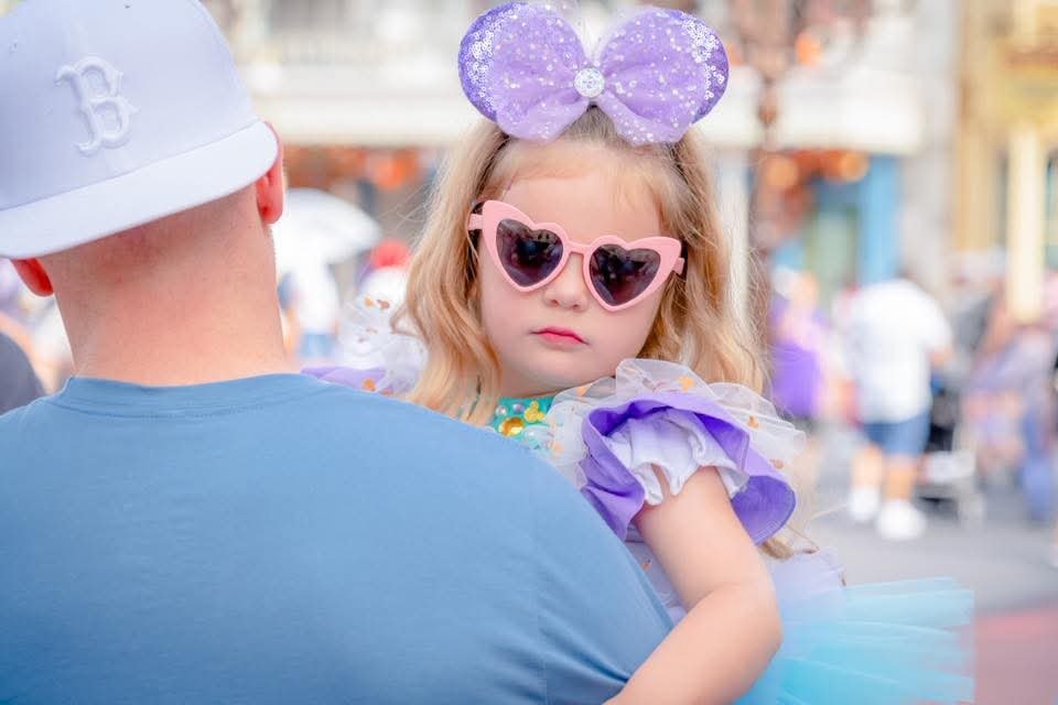 Toddler in heart-shaped sunglasses and a purple Minnie bow being carried on Main Street U.S.A. at Magic Kingdom